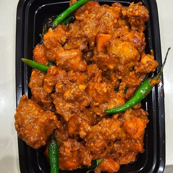 Close-up of orange-red chicken pieces with green chilies in a black takeout container.