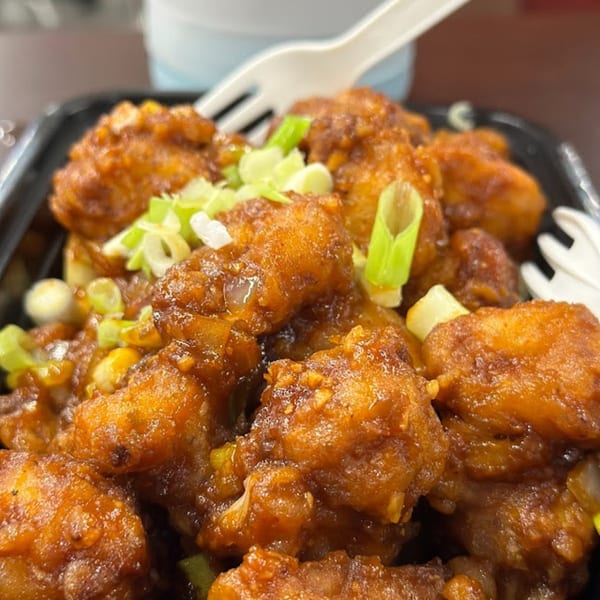 Close-up of crispy orange chicken pieces garnished with fresh green onions in a black takeout container.