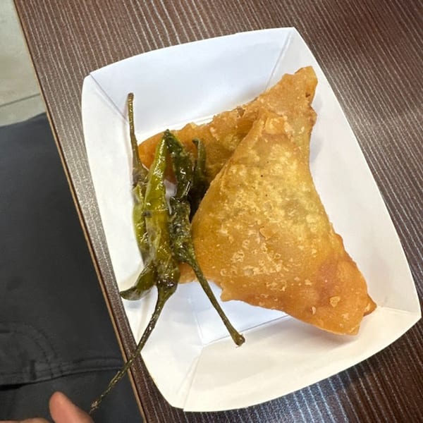 Two golden-brown samosas and several green fried chilies in a white paper tray on a wooden table.